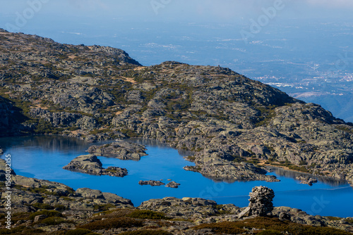 High-angle view of a vivid blue mountain lake set in the granite plateau of Serra da Estrela Natural Park. A small stone cairn stands in the foreground, while rough rock ridges surround the quiet wate