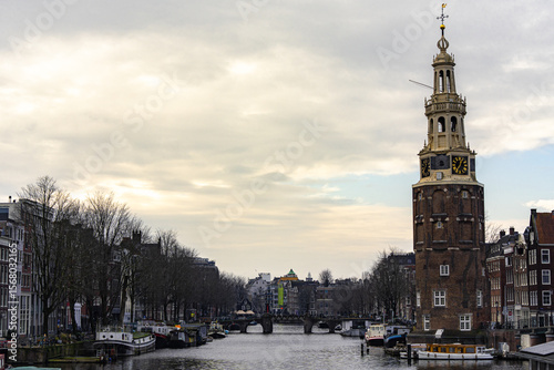 Brick clock-tower “Montelbaanstoren” stands on the edge of the Oude Schans canal. Houseboats line the water under a quiet winter sky, and arched bridges close the view in the distance. 