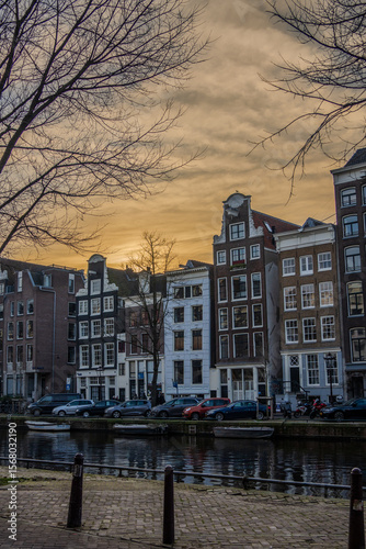 Vertical shot of classic Amsterdam canal houses at sunset. Narrow gabled facades stand along the water while bare winter trees frame a soft orange sky. 