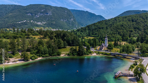 Drone photo of Lake Bohinj in Triglav National Park. The stone bridge and the Church of St John the Baptist sit beside clear turquoise water, with rowboats drifting below forested Julian-Alps slopes. 