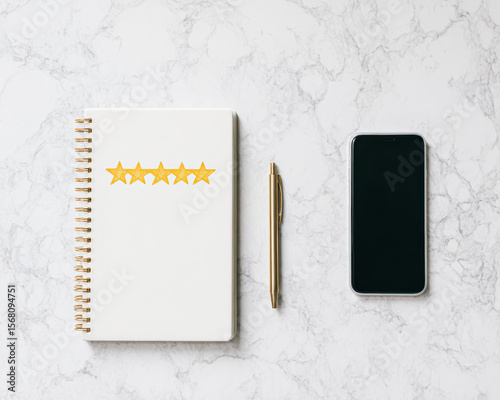 A spiral notebook with golden five stars, a gold pen, and a smartphone arranged neatly on a white marble surface.