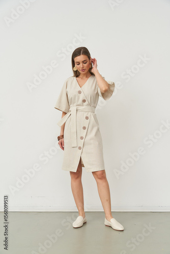 Elegant woman in a light buttoned dress stands with a relaxed pose, hand touching ear, wearing light loafers. Her other hand rests on her hip. White backdrop enhances minimalistic style