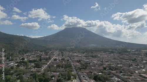 4K aerial footage flying toward Volcán de Agua from Cerro de la Cruz viewpoint above Antigua, Guatemala. The camera reveals the colonial city and majestic volcano under a blue sky.