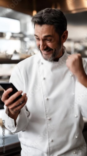 A happy chef in a professional kitchen happily reacting to a message that arrived on his smartphone. Dressed in a white chef's uniform, smiling broadly. Culinary success, restaurant business          