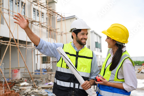 Young attractive construction man and woman in vests with helmets working on the under-construction building site. Home building project. Engineer foreman discusses with a coworker at workplace.