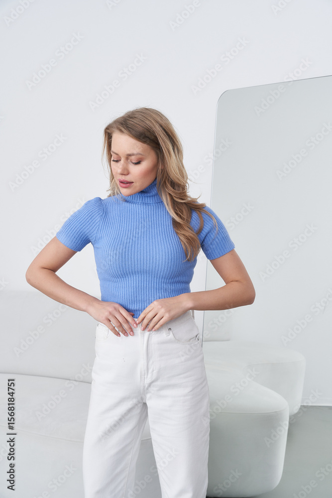 Woman with long hair in a blue ribbed turtleneck and white pants stands in a modern, minimalistic room, adjusting her waistband with a neutral expression and relaxed posture Woman with long hair in a blue ribbed turtleneck and white pants stands in a modern, minimalistic room, adjusting her waistband with a neutral expression and relaxed posture