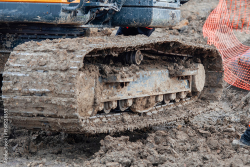 Construction worker  in bright safety gear working hard using shovel to clean tracks of his heavy machinery excavator on muddy site