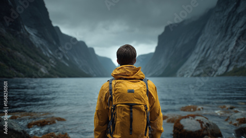 A person with a yellow jacket and backpack looking at a lake surrounded by mountains on a cloudy day