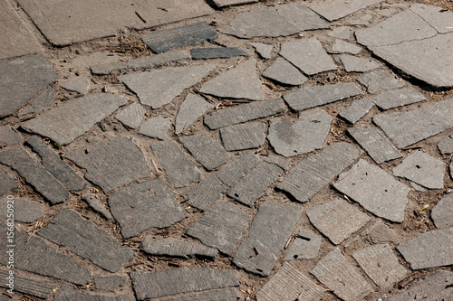 Old stone-paved path made of irregular granite slabs. The surface is uneven, with debris between the stones and small depressions from wear and age.