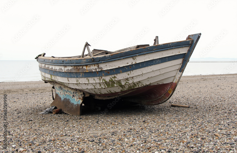 Naklejka premium Abandoned fishing boat on pebbly beach under overcast sky.
