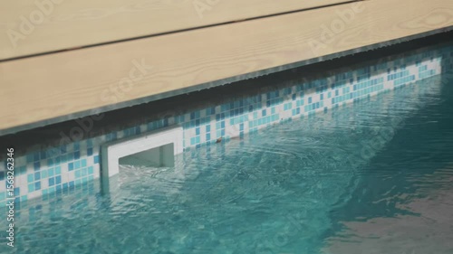 A pool skimmer is built into a tiled wall beneath a wooden deck. Rippling blue water indicates the operation of the filtration system

