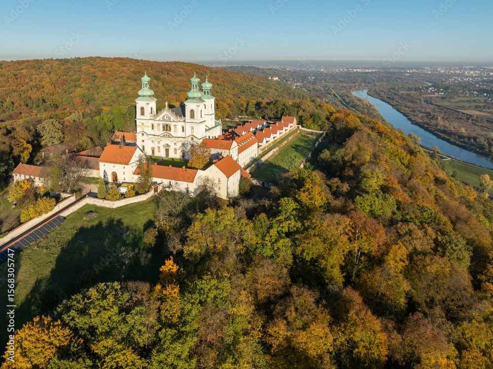 Obraz premium Active Camaldolese monastery and baroque church in a wood on the hill in Bielany, Krakow, Poland. Aerial view in sunset light in fall with Vistula River and far view of Cracow city in the background