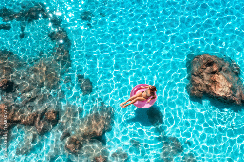 Aerial view of a young woman swimming with pink swim ring in blue sea at sunset in summer. Tropical landscape with girl, clear water, stones, rocks, sandy beach. Top view. Lefkada island, Greece