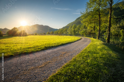 Sun setting behind mountains illuminating gravel road winding through green grass field next to a river with trees and forest in the background during a beautiful spring evening in Slovenia. Sunset