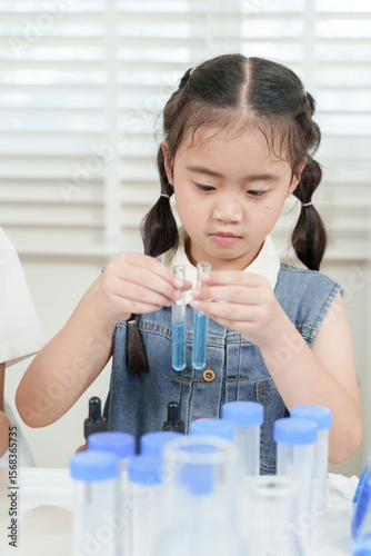 Asian elementary girl student mixing chemicals with test tubes during hands-on science experiment in modern classroom setting surrounded by blue liquid containers and pipettes