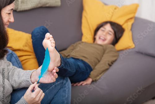 Fotografie Mother tickles son's bare foot with feather while he laughs on the sofa