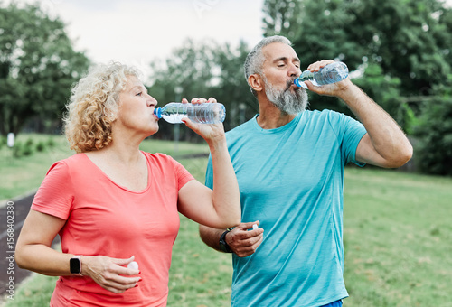 Smiling active mature mid aged couple jogging exercising and having fun and laughing together taking a break drinking water  in the park