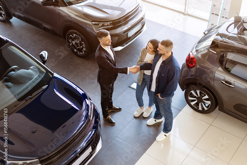 Bild auf Leinwand Dealer shaking hands with young couple in car showroom from aerial view