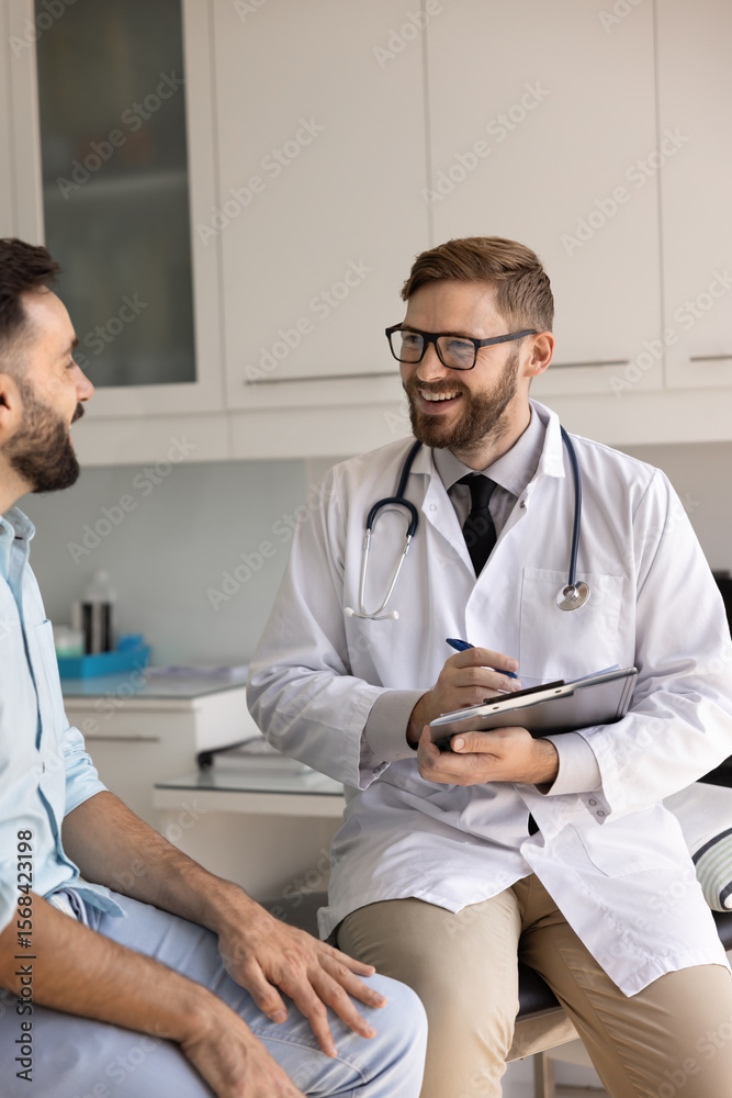 Obraz premium Positive young physician man asking patient about healthcare, disorder symptoms, wellness, writing notes on pad, filling up survey, smiling, laughing, sitting on medical couch. Vertical shot