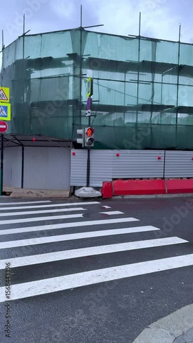 Construction site with scaffolding covered in green mesh, traffic lights visible, and pedestrian crosswalk in foreground, illustrating urban development and infrastructure progress