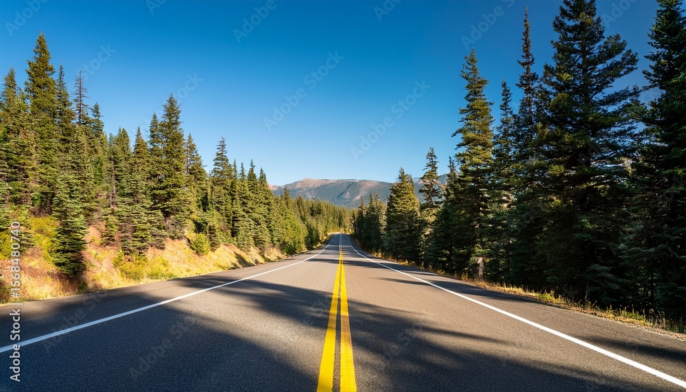 Naklejka premium forest highway lined with tall pines and mountainous backdrop featuring crisp yellow road lines and strong natural light under clear blue sky midday drive scene
