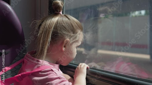 Adorable child girl looking out the train window watching the train station outside waiting for departure wearing a pink shirt and carrying a small backpack Child looking out the window at the train.