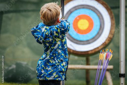 A young boy aims a bow at a colorful archery target, focusing intently while holding the string back, wearing a vibrant blue patterned jacket.