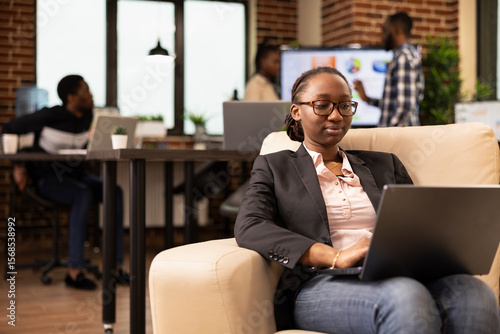 Black female company advisor using laptop, seated on couch, analyzing marketing strategy and compiling reports. African american woman manager comfortably working on business project in startup office