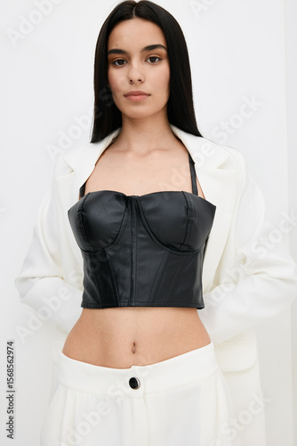 Young woman with long dark hair wearing black leather bustier and white pants stands confidently against plain background, showcasing modern fashion style and elegance.