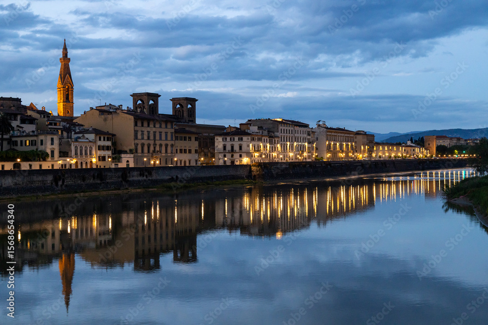 Obraz premium Illuminated buildings along the Arno River reflecting on the water during blue hour in Florence city center, Italy