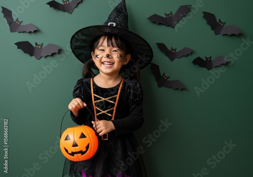 A happy child dressed in a witch costume celebrates Halloween with a pumpkin pail.
