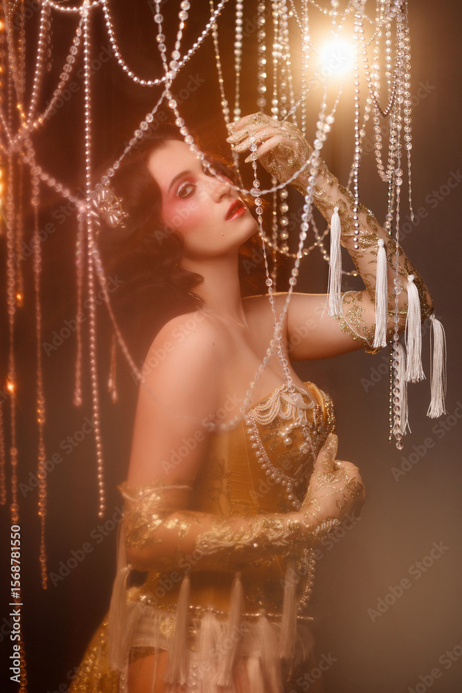 Φωτογραφία Young woman with wavy dark hair, wearing gold vintage corset, posing front view on dramatic stage with hanging bead decor