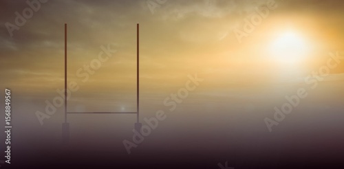 Emerging in mist, H-shaped goalposts standing on open field, sun shining through scattered clouds