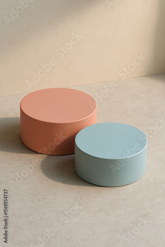 Two cylindrical podiums in dusty rose and sky grey sit atop a raw-textured surface background, top-down oblique, high-angle perspective, dimensional composition