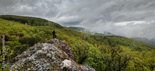 Obraz na płótnie A hiker stands on the rugged edge of Buzgó-kő in the Bükk Mountains, overlooking a vast, mist-covered green valley after spring rain