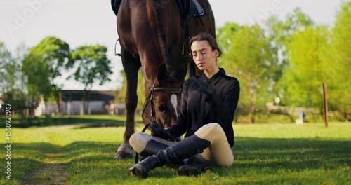 A stylish woman is sitting on the grass next to a horse. She poses looking at the camera