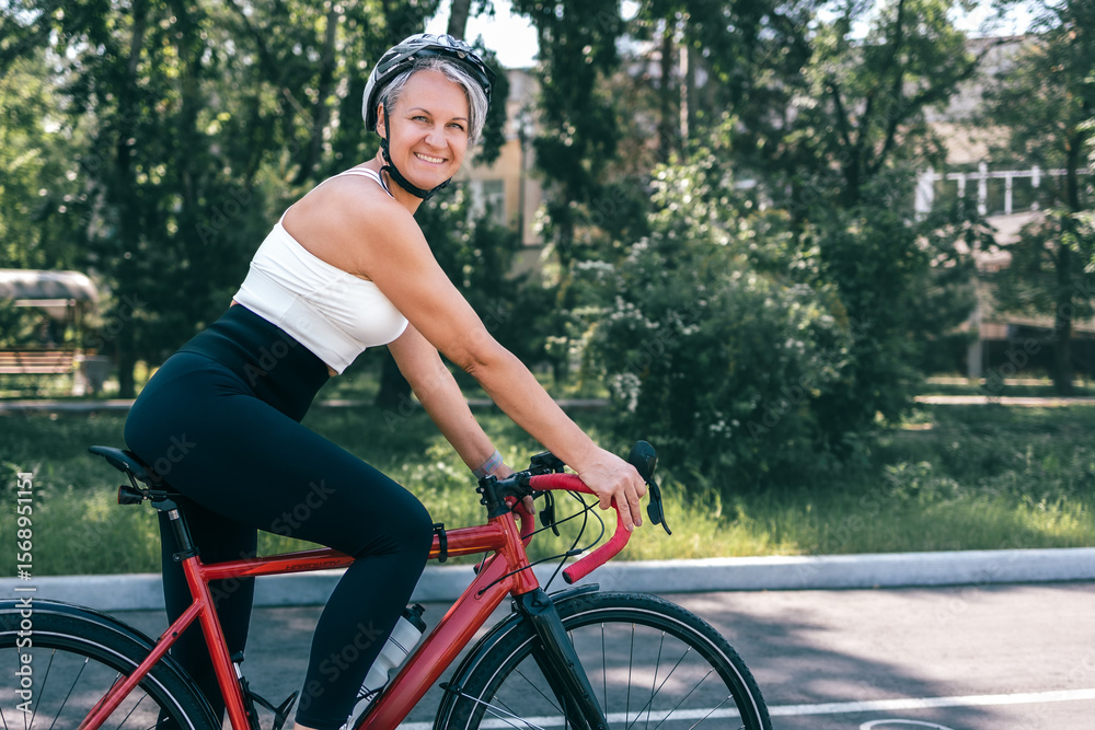 Obraz premium Smiling active senior woman riding red bicycle in park, wearing helmet and sporty outfit