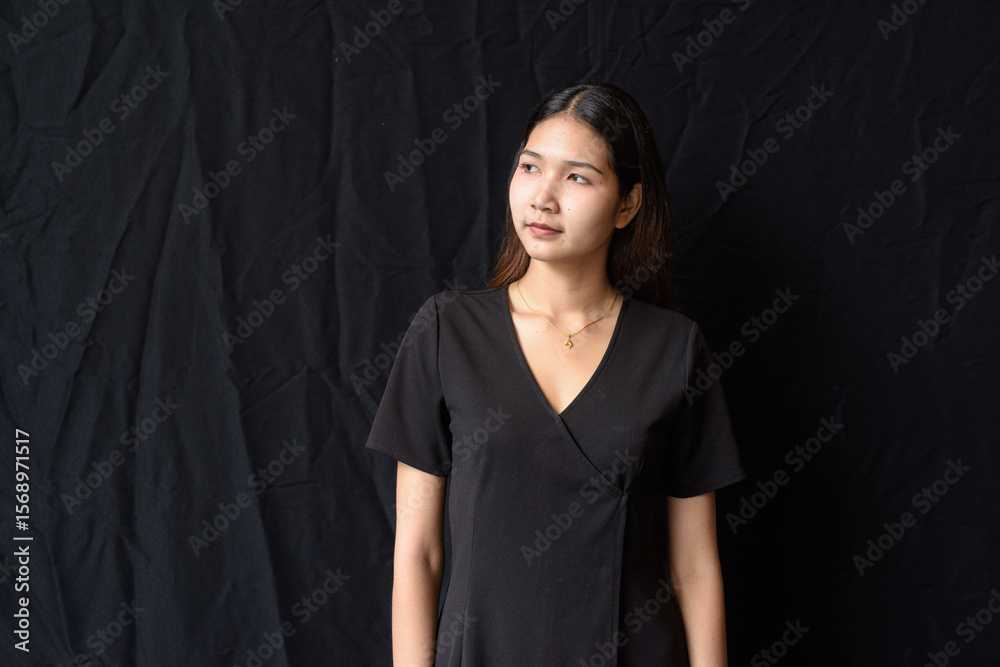 Fototapeta premium Asian woman in black dress posing against black curtain in natural light