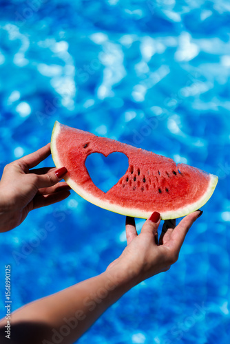 A hand holds a piece of watermelon with a heart-shaped cutout against a swimming pool. The vivid red fruit and symbolic heart shape create a cheerful, romantic summer mood filled with freshness and 