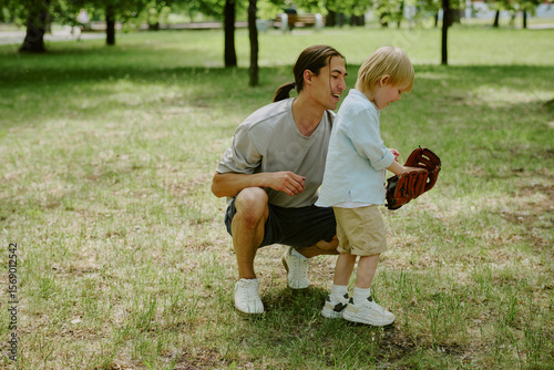 Asian young adult man kneeling beside his child boy teaching him how to use baseball glove in park, both focused on activity, trees and grass visible in background