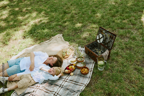 Caucasian middle aged woman and child lying on picnic blanket in park relaxing together near basket with food and drinks, both looking up while enjoying outdoor leisure time