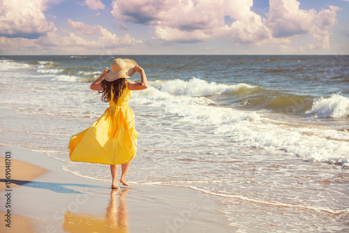 Seascape on a sunny day in summer. Woman on the beach, summertime. Young happy woman walks carefree on the seaside in yellow fluttering dress and sunhat. View from above