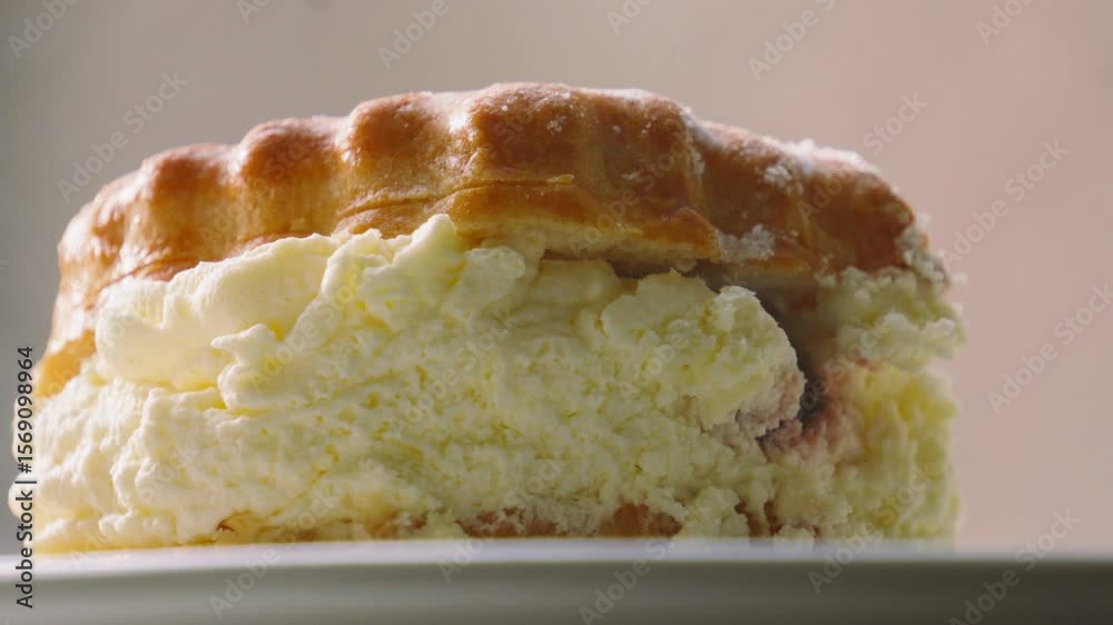 Side View of Sumptuous Traditional Cream and Jam Scone with Light Dusting of Icing Sugar on Top Resting on Plate in Natural Light. British Dessert from Devon and Cornwall. Classic Afternoon Tea. Side View of Sumptuous Traditional Cream and Jam Scone with Light Dusting of Icing Sugar on Top Resting on Plate in Natural Light. British Dessert from Devon and Cornwall. Classic Afternoon Tea.