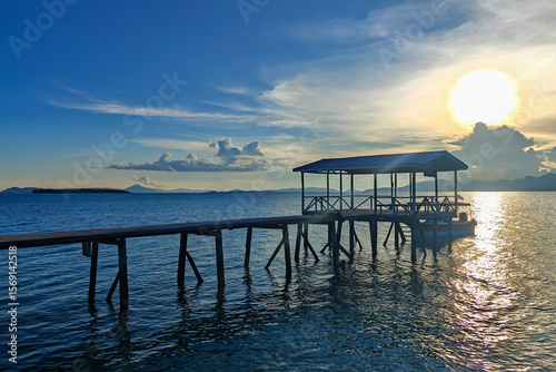 A peaceful wooden jetty at sunset on one of the islands near Semporna, Sabah, Malaysia. A small boat is moored gently on the calm sea as golden light bathes the horizon.