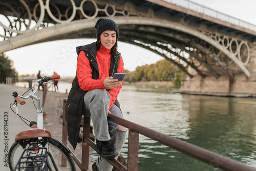 Young woman using mobile phone sitting on railing by bicycle at riverbank