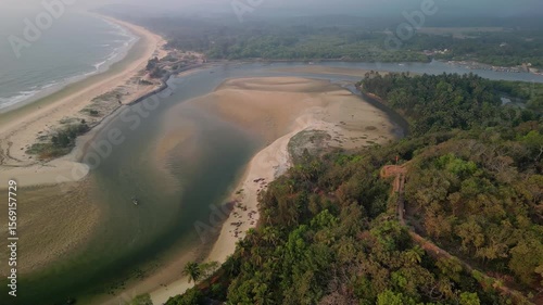 Aerial view shows broad estuary where river meets sea in Goa. Sandbars and vegetation form patterns along shoreline. Watercraft and faint sea haze complete the serene scene