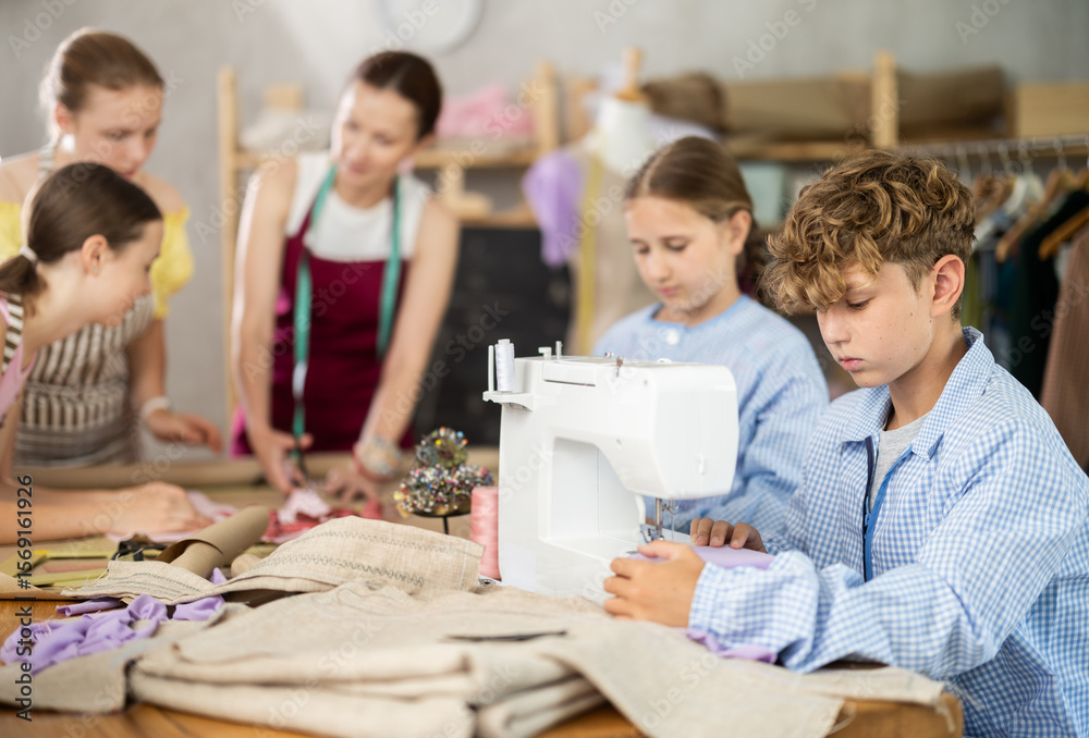Fototapeta premium Teen male student sews product using sewing machine. Pupil are actively cooperating with each other in background, discussing completion of assignment