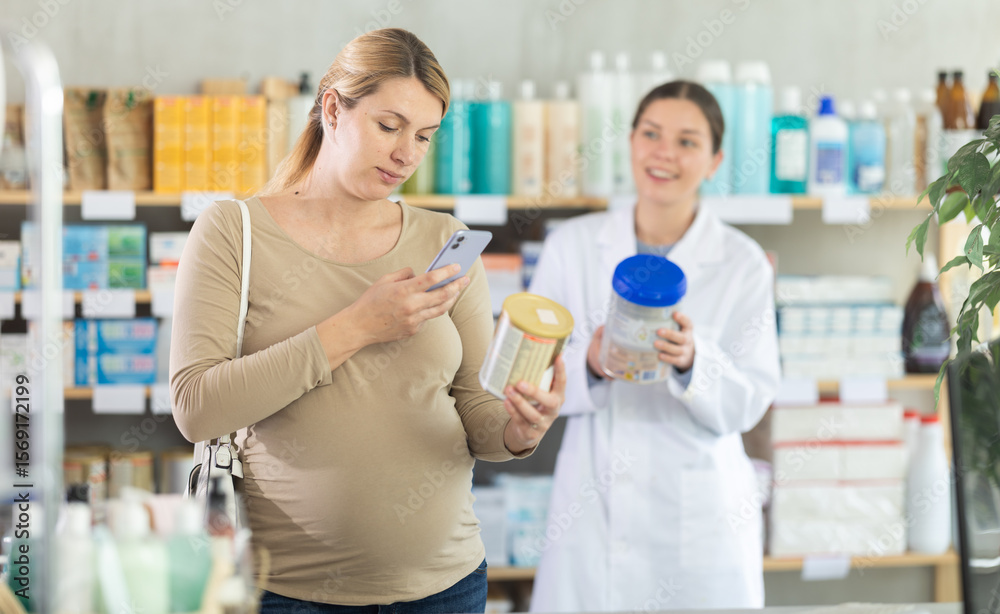 Fototapeta premium Pregnant woman in pharmacy scans QR code on infant formula product, studies product in detail, collects information online. Girl pharmacist offer similar product in background.
