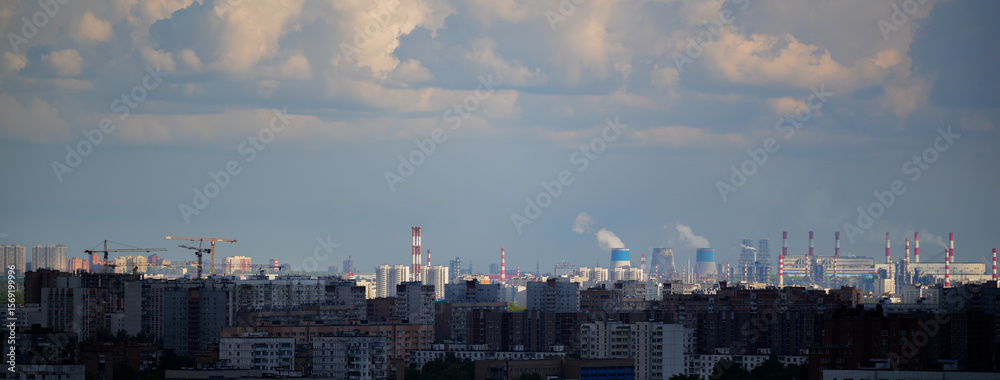 Obraz premium Cityscape showing a mix of residential buildings, construction sites with tower cranes, and a distant industrial zone with chimneys and cooling towers emitting steam. Concept of urban development, cit