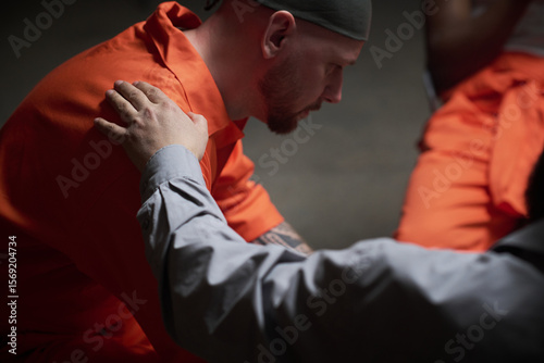 Caucasian man wearing prison uniform sitting with head bowed receiving comforting gesture from therapist during group session in correctional facility